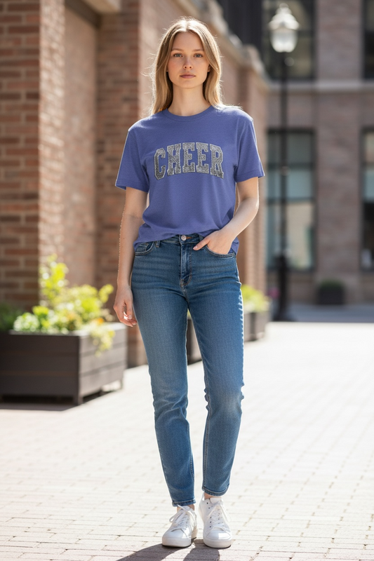 A woman standing on a brick sidewalk wearing a slate blue t-shirt with "CHEER" printed in a distressed athletic font. She is paired with slim-fit blue jeans and white sneakers.
